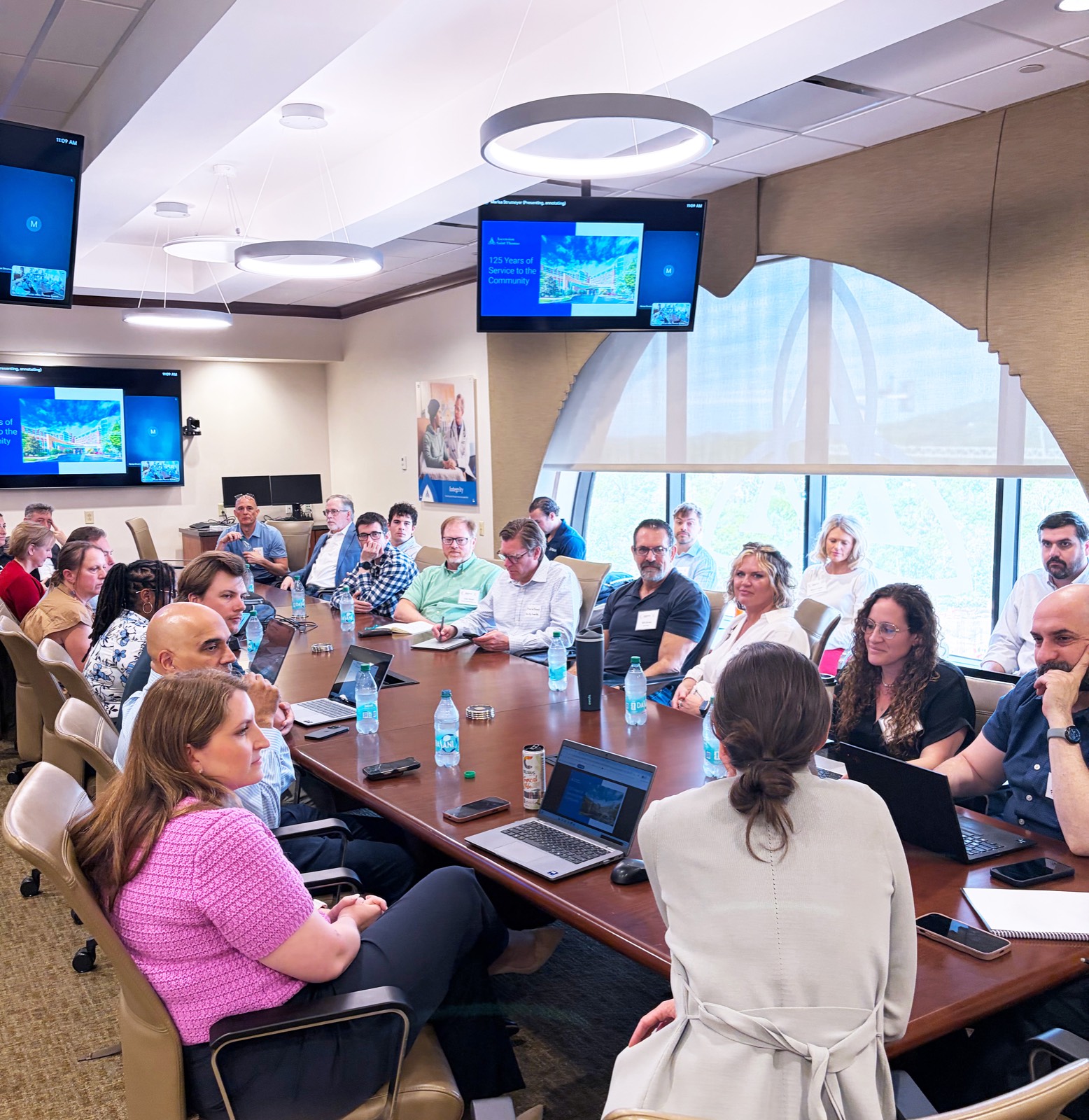Roughly two dozen founders and health system staff seated around a long boardroom table during an immersion visit, with a presentation on screen behind them.